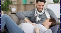 A man and a pregnant woman sitting together on a couch, both smiling warmly and hoping to have twin babies